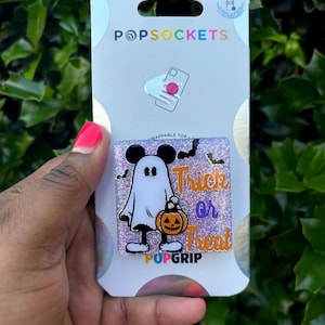 May include: A white ghost with Mickey Mouse ears holding a pumpkin bucket with the text "Trick or Treat" on a purple glitter background. The background is a white card with the text "PopSockets" and "PopGrip".