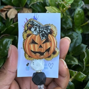May include: A Halloween-themed badge reel featuring a smiling jack-o'-lantern with a black and white bow, surrounded by sunflowers. The reel has a white and black bead design. The card reads "Thank you for supporting us".