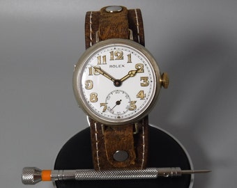 May include: A vintage Rolex wristwatch with a white face and black hands. The watch has gold Arabic numerals and a brown leather strap.