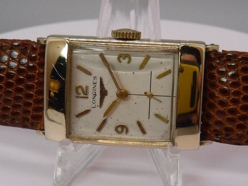 May include: A gold-tone Longines wristwatch with a white face and black hands. The watch has a brown leather band.