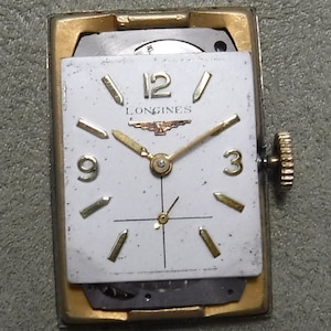 May include: A gold-toned Longines wristwatch with a white face and black hands. The watch has a square face and a gold-toned band.