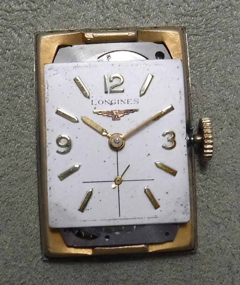 May include: A gold-toned Longines wristwatch with a white face and black hands. The watch has a square face and a gold-toned band.