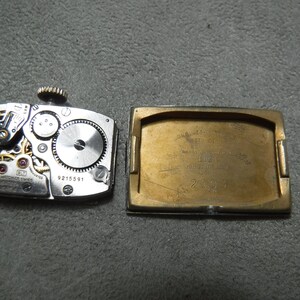 May include: The back of a gold-toned wristwatch with a silver-toned face. The watch face is visible through the open back of the watch case. The watch case has the text "ONWARD AUTOMATIC 17 JEWELS SWISS MADE" and "LIEGI" with a number "20924". The watch movement has the number "9215591".