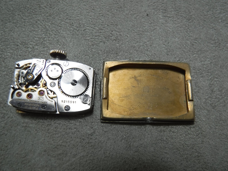 May include: The back of a gold-toned wristwatch with a silver-toned face. The watch face is visible through the open back of the watch case. The watch case has the text "ONWARD AUTOMATIC 17 JEWELS SWISS MADE" and "LIEGI" with a number "20924". The watch movement has the number "9215591".