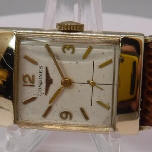 May include: A gold-tone Longines wristwatch with a white face and black hands. The watch has a square face and a brown leather band.