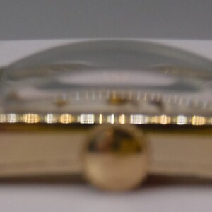 May include: A gold-toned wristwatch with a brown leather band. The watch face is round and has a white background with black markings.