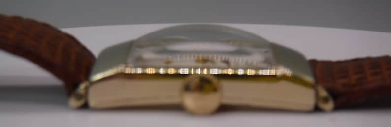 May include: A gold-toned wristwatch with a brown leather band. The watch face is round and has a white background with black markings.
