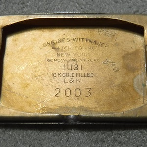 May include: Close-up of the back of a gold-toned watch case. The case is engraved with the text "LONGINES-WITTNAUER WATCH CO INC. NEW YORK GENEVA MONTREAL L131 10 K GOLD FILLED L&K 2003".