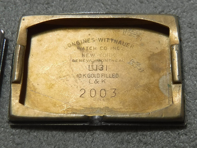 May include: Close-up of the back of a gold-toned watch case. The case is engraved with the text "LONGINES-WITTNAUER WATCH CO INC. NEW YORK GENEVA MONTREAL L131 10 K GOLD FILLED L&K 2003".