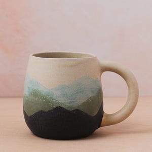 May include: A ceramic mug with a mountain landscape design. The mug has a cream-coloured top, with a gradient of blue, green, and black at the base. The handle is a rounded, loop shape. Suitable for hot or cold drinks.