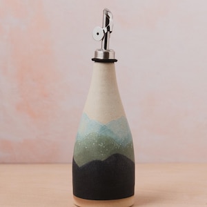 May include: Ceramic oil bottle with a stainless steel spout. The bottle features a landscape design with blue, green, and black hues. The base is a natural beige colour.