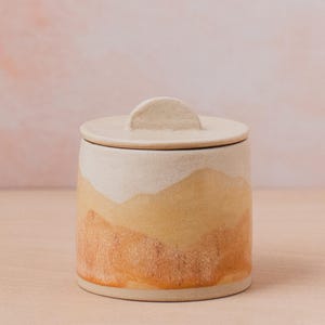 May include: A ceramic jar with a lid featuring a rounded handle. The jar has a white base with a yellow and orange gradient design.