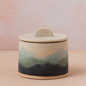 May include: A small ceramic jar with a lid, featuring a hand-painted landscape design in shades of blue, green, and black. The jar has a rounded shape and a smooth, matte finish.
