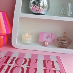 May include: A white arched shelf containing a silver disco ball planter with a green plant, a lit candle, a pink card, a small pink billiard ball, and a glass jar. In the foreground, a pink and white striped sign reads SHOP IT SHOP IT Real good.