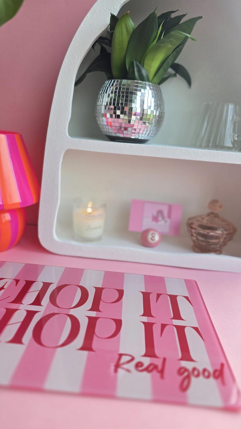 May include: A white arched shelf containing a silver disco ball planter with a green plant, a lit candle, a pink card, a small pink billiard ball, and a glass jar. In the foreground, a pink and white striped sign reads SHOP IT SHOP IT Real good.