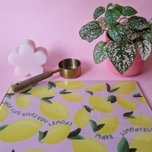 May include: A rectangular glass cutting board with a pink background, yellow lemon illustrations, green leaves, and the text "WHEN LIFE GIVES YOU LEMONS MAKE LIMONCELLO". A pink flower-shaped candle, a gold measuring scoop, and a potted plant are nearby.