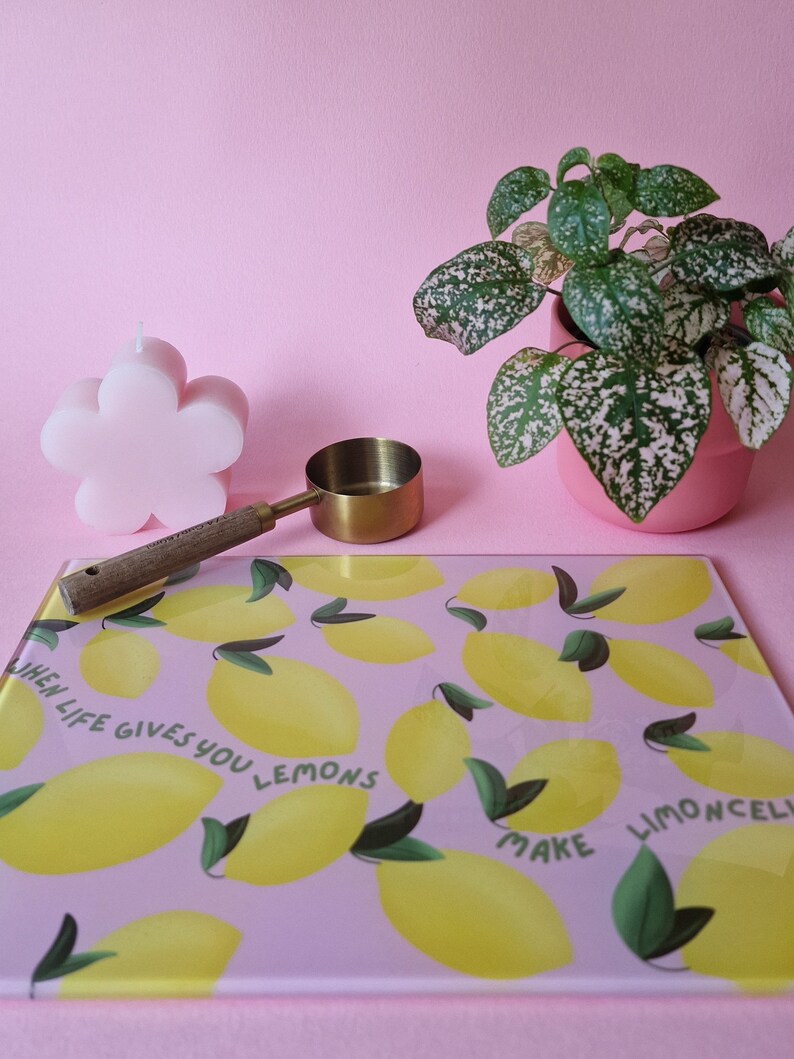 May include: A rectangular glass cutting board with a pink background, yellow lemon illustrations, green leaves, and the text "WHEN LIFE GIVES YOU LEMONS MAKE LIMONCELLO". A pink flower-shaped candle, a gold measuring scoop, and a potted plant are nearby.