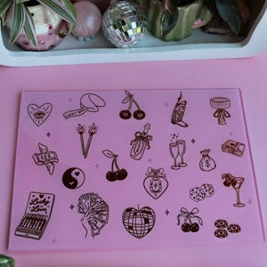 May include: A rectangular pink glass cutting board featuring a variety of dark brown line art illustrations including cherries, a disco ball, dice, a yin-yang symbol, money, and a matchbook labeled Lucky in Love, paired with a pink-bladed knife.