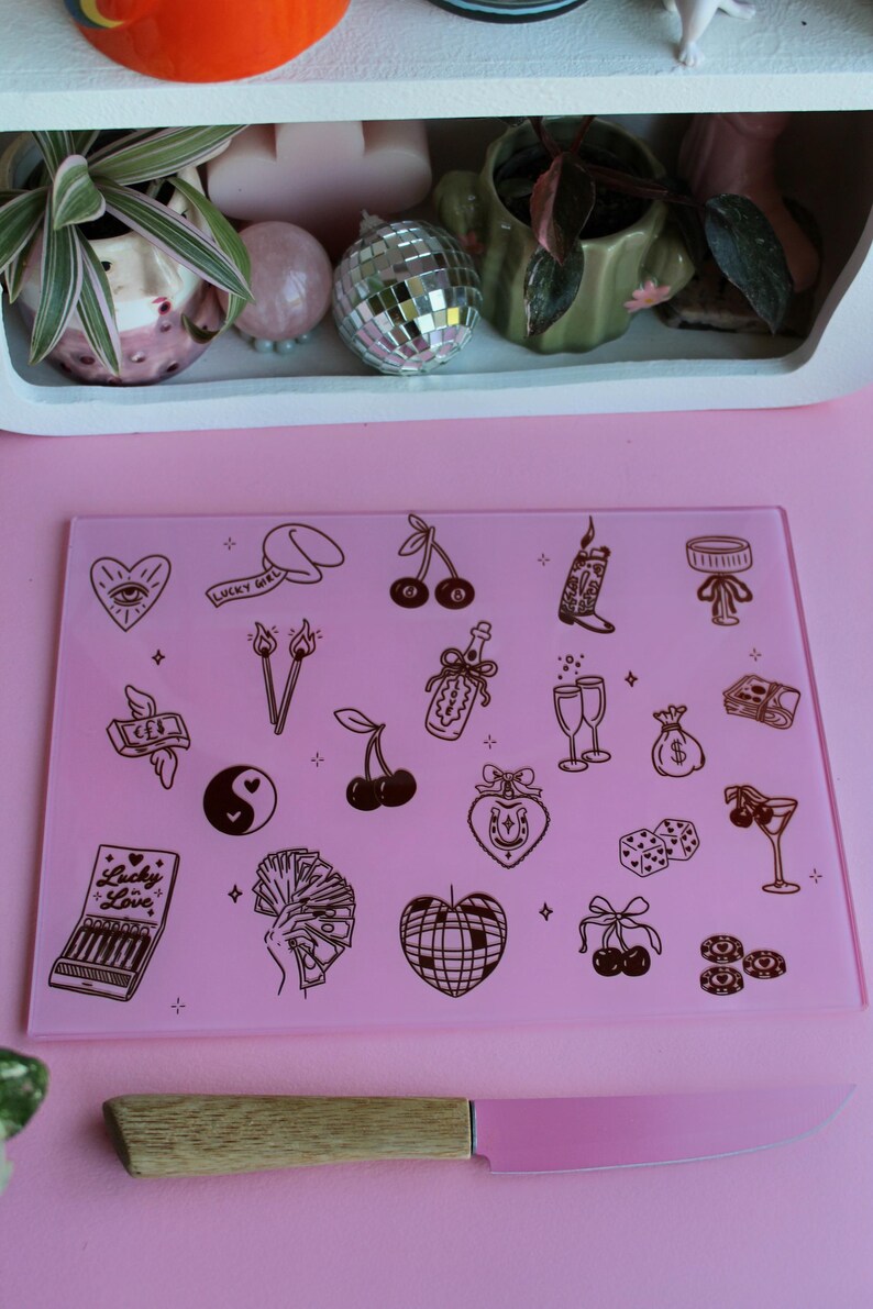 May include: A rectangular pink glass cutting board featuring a variety of dark brown line art illustrations including cherries, a disco ball, dice, a yin-yang symbol, money, and a matchbook labeled Lucky in Love, paired with a pink-bladed knife.