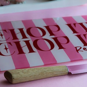 May include: A rectangular glass cutting board with alternating vertical pink and white stripes and the red text CHOP IT CHOP IT, paired with a kitchen knife featuring a light-colored wooden handle and a metallic blade resting in the foreground.