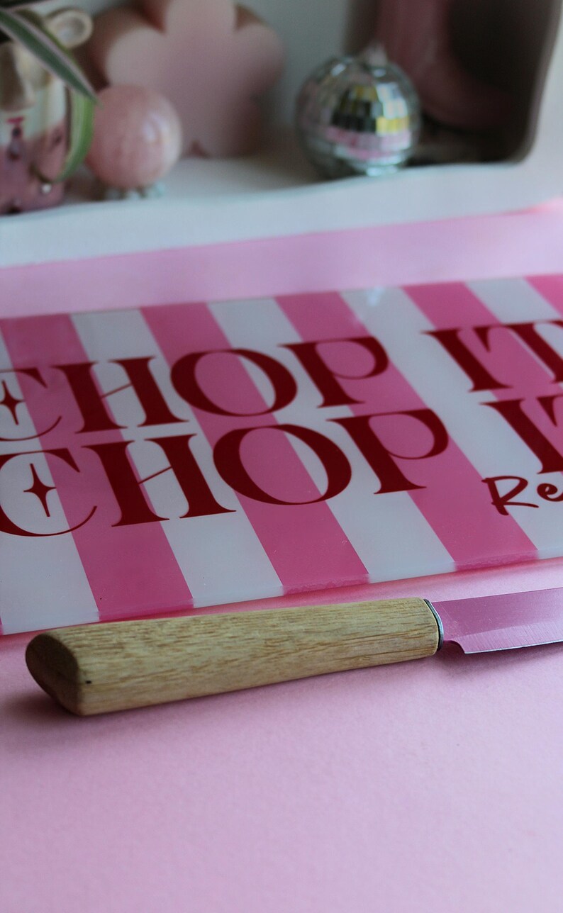 May include: A rectangular glass cutting board with alternating vertical pink and white stripes and the red text CHOP IT CHOP IT, paired with a kitchen knife featuring a light-colored wooden handle and a metallic blade resting in the foreground.