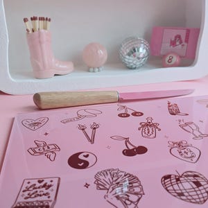May include: A rectangular pink glass cutting board featuring dark brown line art illustrations of cherries, a yin-yang symbol, a hand holding cash, a disco ball heart, and a matchbox labeled Lucky Love, accompanied by a knife with a wooden handle.