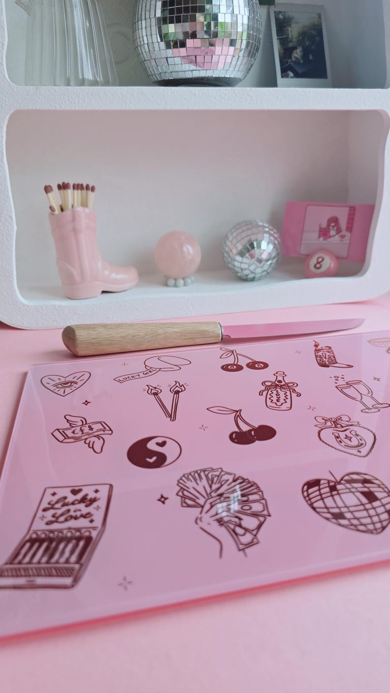May include: A rectangular pink glass cutting board featuring dark brown line art illustrations of cherries, a yin-yang symbol, a hand holding cash, a disco ball heart, and a matchbox labeled Lucky Love, accompanied by a knife with a wooden handle.