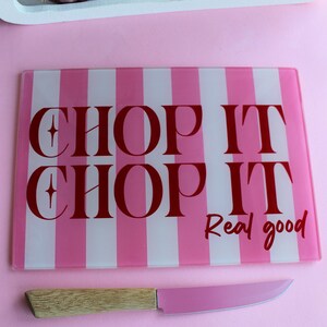 May include: A rectangular glass cutting board with vertical pink and white stripes and the red text "CHOP IT CHOP IT Real good" paired with a knife featuring a light wood handle and a pink blade, positioned on a solid pink surface.