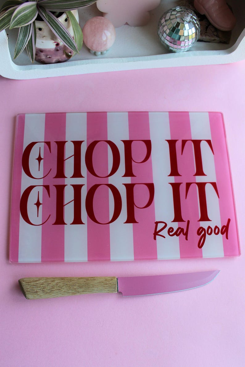 May include: A rectangular glass cutting board with vertical pink and white stripes and the red text "CHOP IT CHOP IT Real good" paired with a knife featuring a light wood handle and a pink blade, positioned on a solid pink surface.