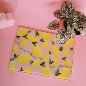 May include: A rectangular glass cutting board featuring a repeating pattern of yellow lemons with green leaves on a light pink background. The text "WHEN LIFE GIVES YOU LEMONS MAKE LIMONCELLO!" is printed in dark green letters across the surface.