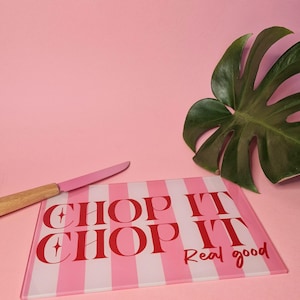 May include: A rectangular glass cutting board featuring a pink and white vertical striped pattern with the red text "CHOP IT CHOP IT Real good" printed on the surface, accompanied by a knife with a pink blade and a light brown wooden handle.
