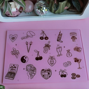 May include: A rectangular pink glass cutting board featuring a variety of dark brown line art illustrations including cherries, a disco ball, dice, a yin-yang symbol, money, and a matchbook labeled Lucky in Love, paired with a pink-bladed knife.