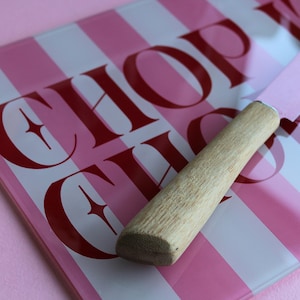 May include: A rectangular glass cutting board with vertical pink and white stripes and the text CHOP CHOP in dark red lettering. A knife with a light pink blade and a light-colored wooden handle rests diagonally across the cutting board surface.
