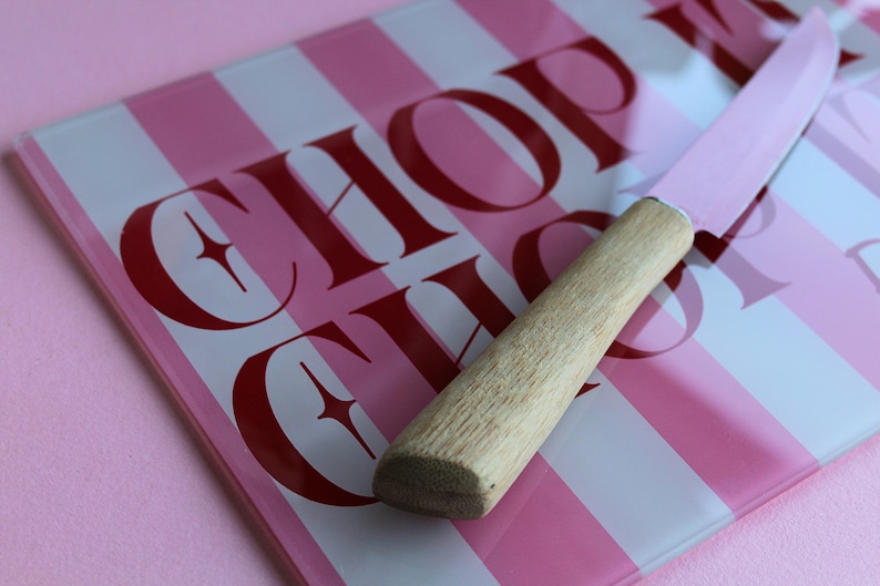 May include: A rectangular glass cutting board with vertical pink and white stripes and the text CHOP CHOP in dark red lettering. A knife with a light pink blade and a light-colored wooden handle rests diagonally across the cutting board surface.