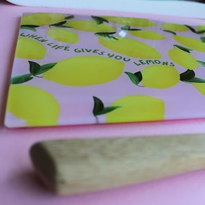May include: A rectangular glass cutting board with a light pink background featuring a pattern of yellow lemons with green leaves. The text "WHEN LIFE GIVES YOU LEMONS" is printed in dark green letters across the center of the board.