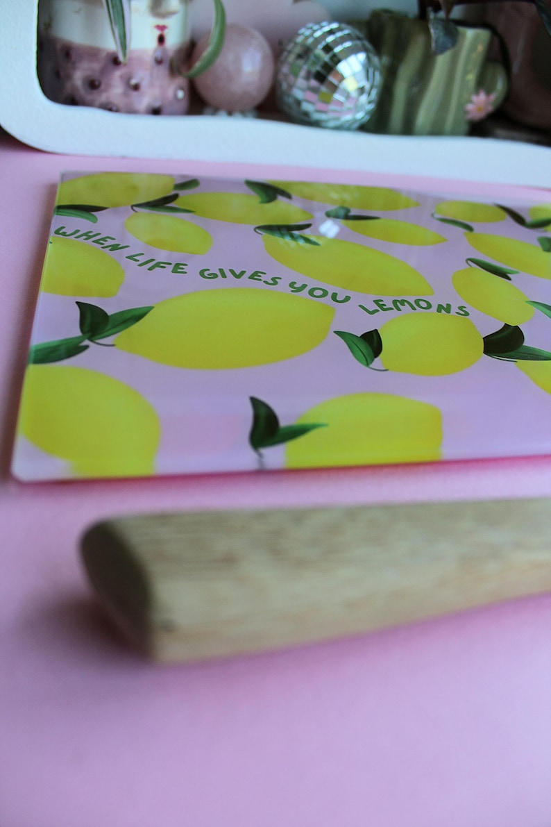 May include: A rectangular glass cutting board with a light pink background featuring a pattern of yellow lemons with green leaves. The text "WHEN LIFE GIVES YOU LEMONS" is printed in dark green letters across the center of the board.