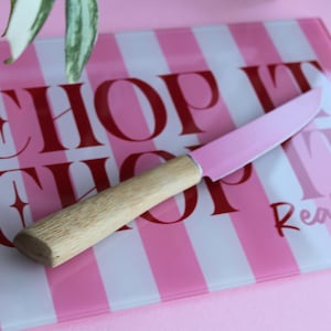 May include: A rectangular glass cutting board with pink and white vertical stripes and the red text "CHOP IT" and "Real good". A kitchen knife with a light wood handle and a pink blade rests diagonally across the center of the cutting board.