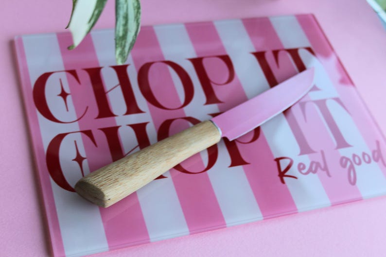 May include: A rectangular glass cutting board with pink and white vertical stripes and the red text "CHOP IT" and "Real good". A kitchen knife with a light wood handle and a pink blade rests diagonally across the center of the cutting board.