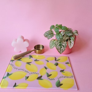 May include: A rectangular glass cutting board with a light purple background, yellow lemon illustrations, and the text "WHEN LIFE GIVES YOU LEMONS MAKE LIMONCELLO!". Also includes a flower-shaped pink candle, a gold measuring scoop, and a potted plant.