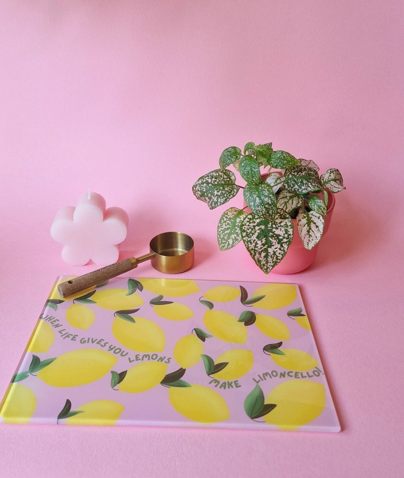 May include: A rectangular glass cutting board with a light purple background, yellow lemon illustrations, and the text "WHEN LIFE GIVES YOU LEMONS MAKE LIMONCELLO!". Also includes a flower-shaped pink candle, a gold measuring scoop, and a potted plant.