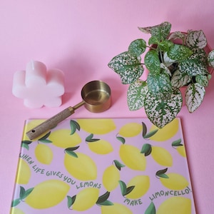 May include: A rectangular glass cutting board with a pink background, yellow lemon illustrations, green leaves, and the text "WHEN LIFE GIVES YOU LEMONS MAKE LIMONCELLO!" alongside a brass measuring scoop with a wooden handle and a pink flower-shaped candle.
