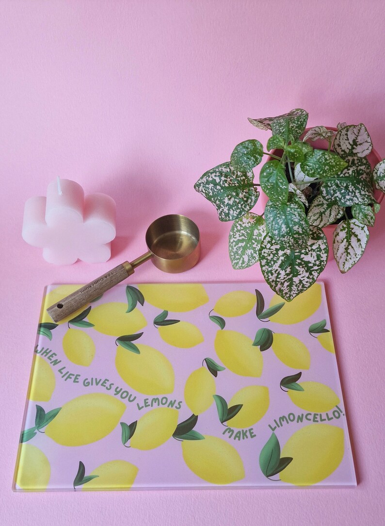 May include: A rectangular glass cutting board with a pink background, yellow lemon illustrations, green leaves, and the text "WHEN LIFE GIVES YOU LEMONS MAKE LIMONCELLO!" alongside a brass measuring scoop with a wooden handle and a pink flower-shaped candle.