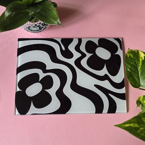 May include: A white and black glass chopping board with a wavy pattern and two black flowers. The flowers have white centres.