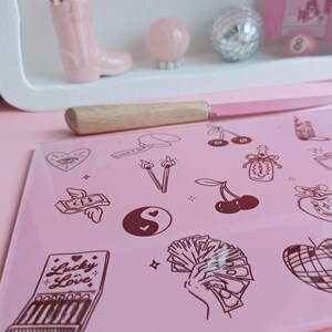 May include: A rectangular pink glass cutting board featuring dark brown line art illustrations including a matchbook labeled Lucky in Love, a hand holding cash, cherries, a yin-yang symbol, a heart with an eye, and a disco ball, paired with a knife.