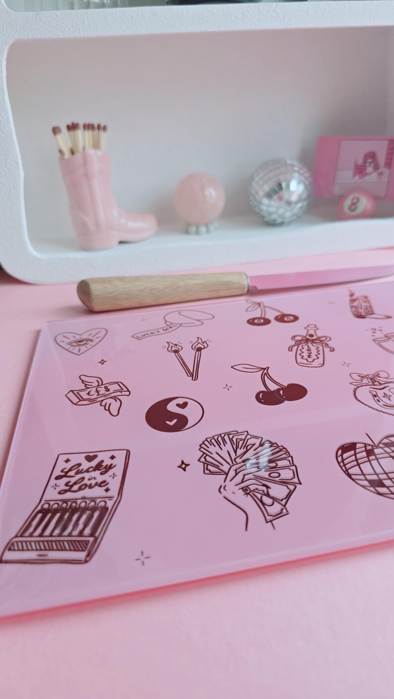 May include: A rectangular pink glass cutting board featuring dark brown line art illustrations including a matchbook labeled Lucky in Love, a hand holding cash, cherries, a yin-yang symbol, a heart with an eye, and a disco ball, paired with a knife.