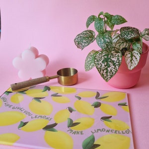 May include: A rectangular glass cutting board with a pink background, yellow lemon illustrations, green leaves, and the text "WHEN LIFE GIVES YOU LEMONS MAKE LIMONCELLO!". A pink flower-shaped candle, a gold measuring scoop, and a potted plant are nearby.
