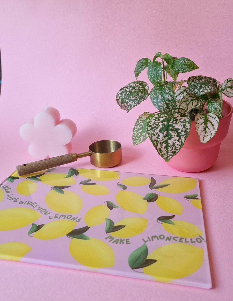 May include: A rectangular glass cutting board with a pink background, yellow lemon illustrations, green leaves, and the text "WHEN LIFE GIVES YOU LEMONS MAKE LIMONCELLO!". A pink flower-shaped candle, a gold measuring scoop, and a potted plant are nearby.