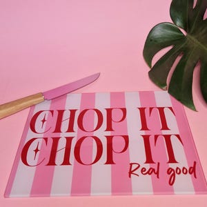 May include: A rectangular glass cutting board featuring vertical light pink and white stripes. The text CHOP IT CHOP IT Real good is printed in bold red lettering across the center. A knife with a pink blade and wooden handle rests nearby.