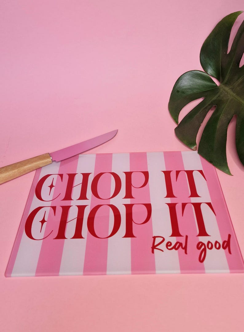 May include: A rectangular glass cutting board featuring vertical light pink and white stripes. The text CHOP IT CHOP IT Real good is printed in bold red lettering across the center. A knife with a pink blade and wooden handle rests nearby.