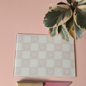 Könnte beinhalten: Ein Schneidebrett aus Glas mit einem rosa-weißen Blumenschachmuster. Ein rosa Messer mit Holzgriff liegt darunter. Der Hintergrund ist einfarbig rosa, mit einer Pflanze in der oberen rechten Ecke.